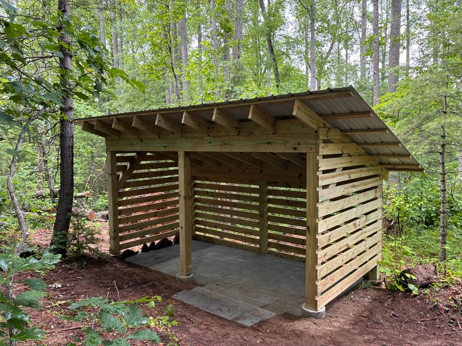 Open-front timber firewood shed with slatted sides, a metal roof and a paver floor